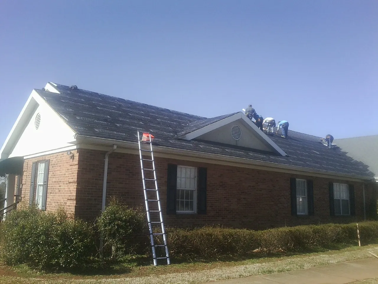 Crew tearing shingles off a brick ranch roof with a ladder set — Emergency Roof Repair work by our crew in Lake Mary